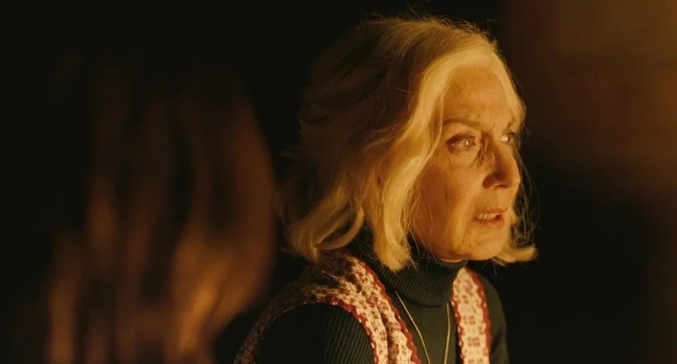 The Skin I Live In (2011) movie still: closeup — A dimly lit indoor space with a dark background and orange light hues. An older woman with blonde…