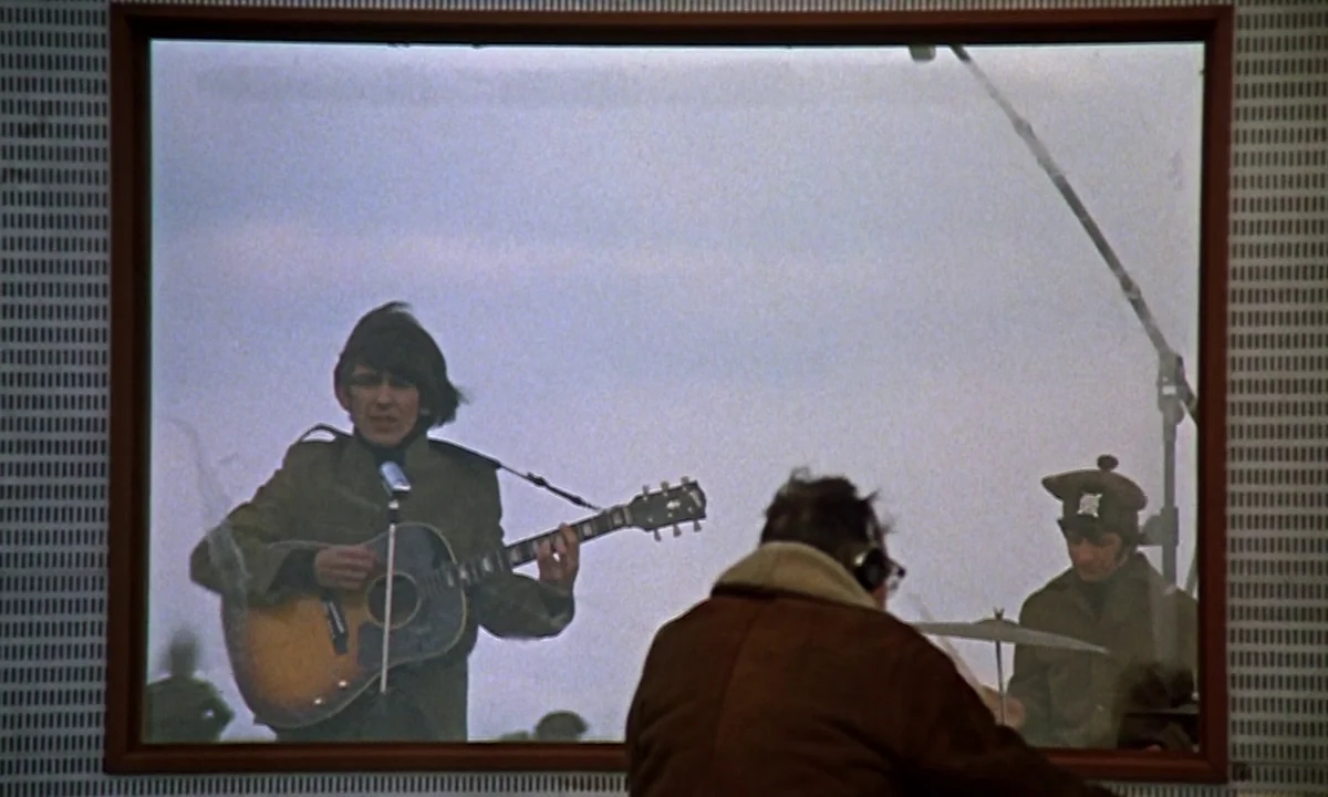 Help! (1965) movie still: medium — Inside a recording studio, a man plays acoustic guitar behind a large window frame. A drummer sits behind him in…