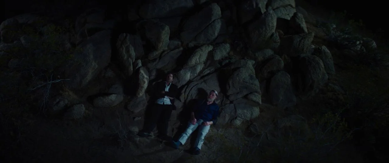 Palm Springs (2020) movie still: wide — A high-angle view shows a rocky, desert-like slope at night with minimal vegetation. Two men lie prone on…