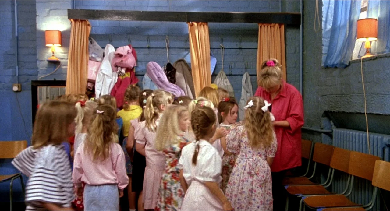 Life Is Sweet (1990) movie still: medium — A blue-walled room with orange curtains acting as dividers features hanging bags and clothes. An adult…