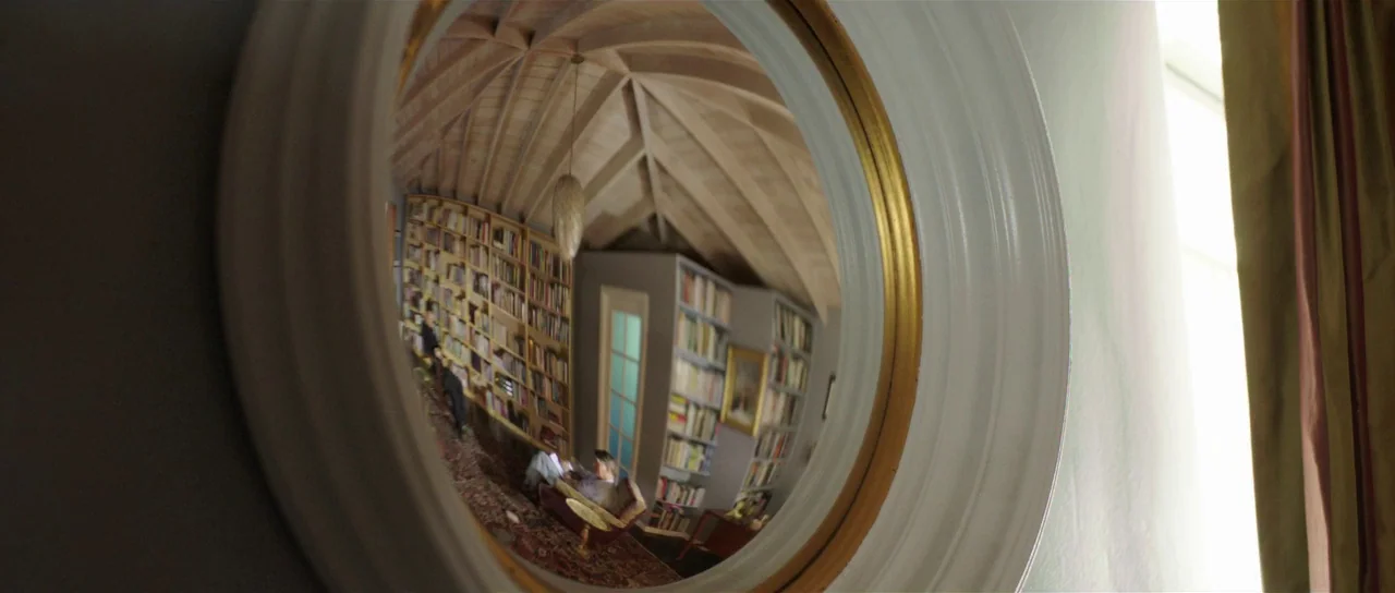 The Canyons (2013) movie still: closeup — A circular mirror reflects a room with a vaulted wood ceiling and tall shelves. Books fill the shelves and…