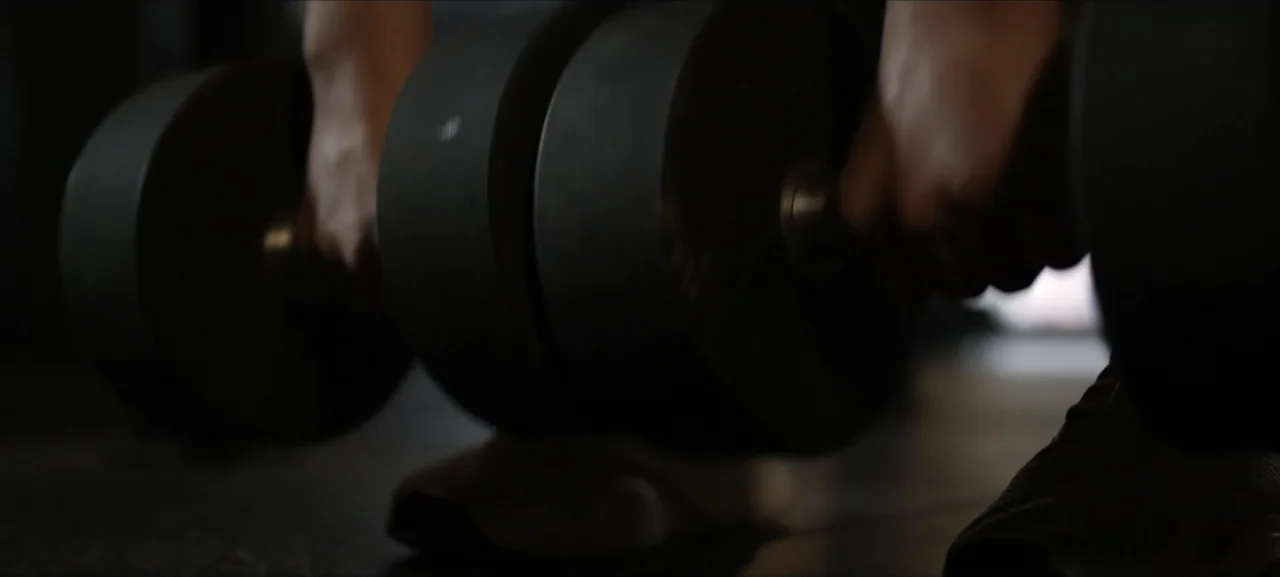 No Woman No Try (2022) movie still: closeup — A dimly lit indoor space features dark rubber flooring for weight training. Two large black metal…