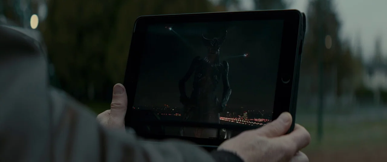 Colossal (2016) movie still: closeup — A pair of human hands hold a tablet outside in a blurred park environment. The tablet screen shows a dark…