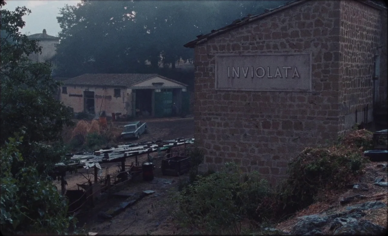 Happy as Lazzaro (2018) movie still: establishing — A rural courtyard features a stone building with the name Inviolata inscribed upon it. A small…