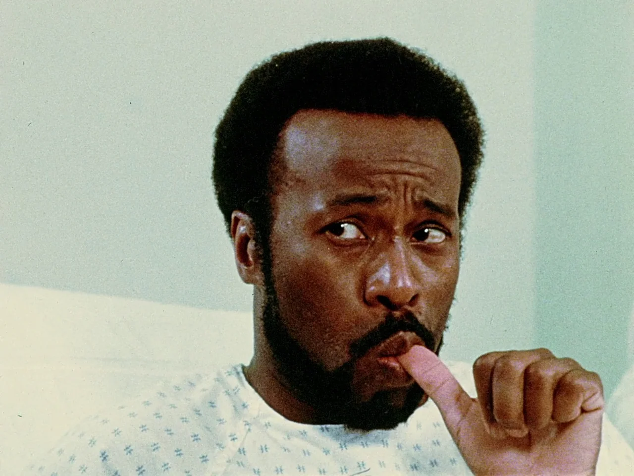 Space is the Place (1974) movie still: closeup — A man lies in a hospital bed against a plain pale wall. He stares off-frame while sucking his thumb.