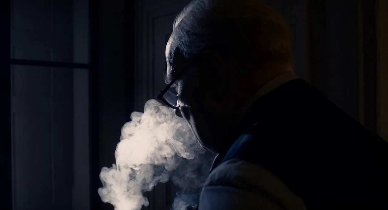 Darkest Hour (2017) movie still: closeup — A dark, dimly lit study with a prominent window frame in the background. An elderly man in a dark vest…