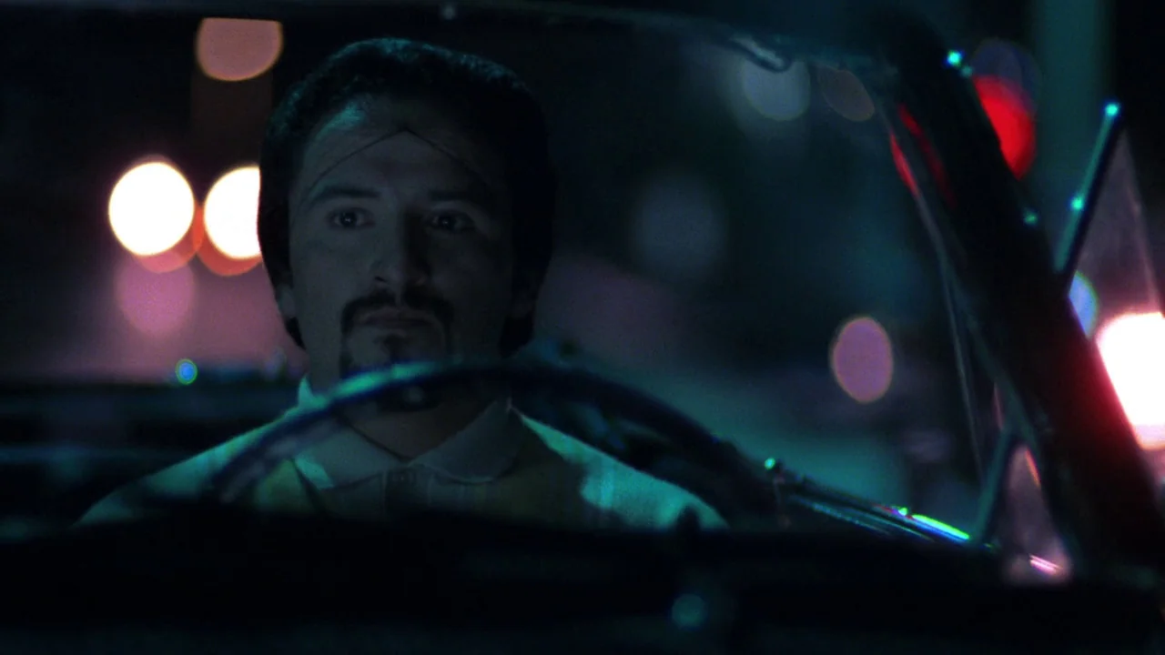 Repo Man (1984) movie still: medium — A man drives a car at night through a city with vibrant neon lights. The interior is dark and illuminated by…