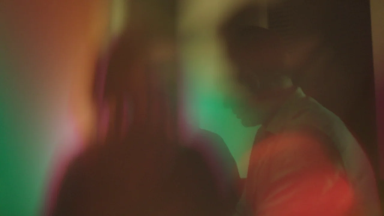 Infinity Pool (2023) movie still: medium — A dimly lit indoor space with heavy red and green light flares overlaying silhouettes. Two individuals…