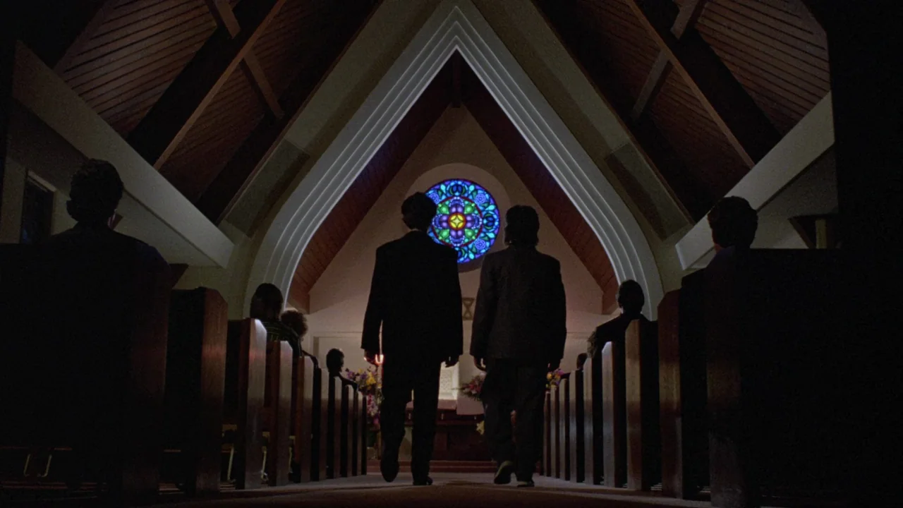 Society (1989) movie still: fullbody — A church interior with wooden pews and a circular stained glass window above the altar. Two men walk down the…