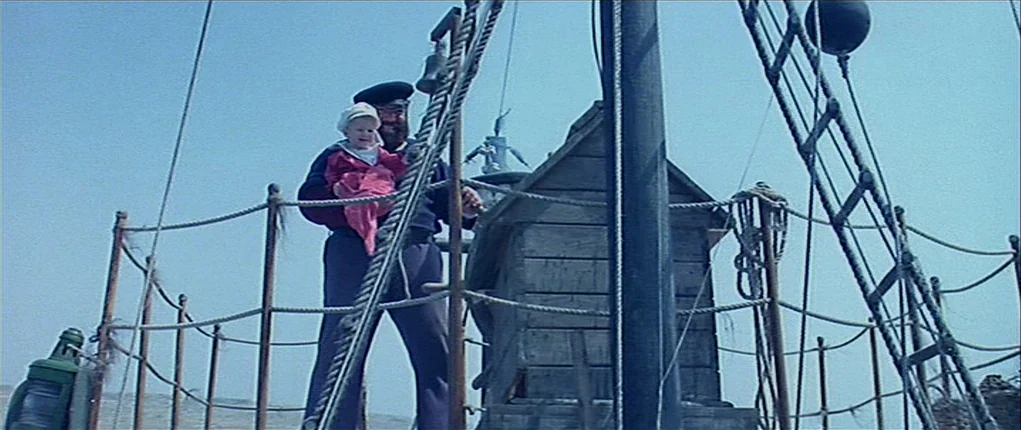 Popeye (1980) movie still: fullbody — The deck of a boat features a wooden structure and ropes under clear skies. A man stands holding a small…