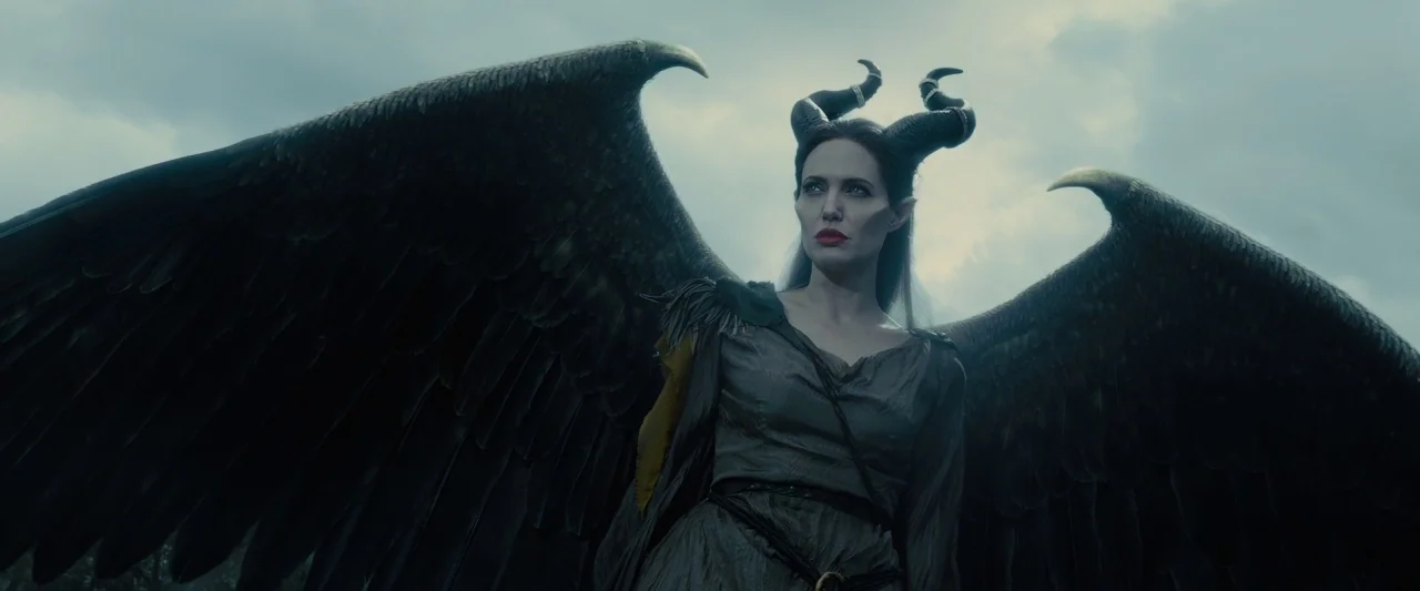 Maleficent (2014) movie still: medium — A figure with massive feathered wings stands against a cloudy, grey sky. The subject wears dark fantasy…