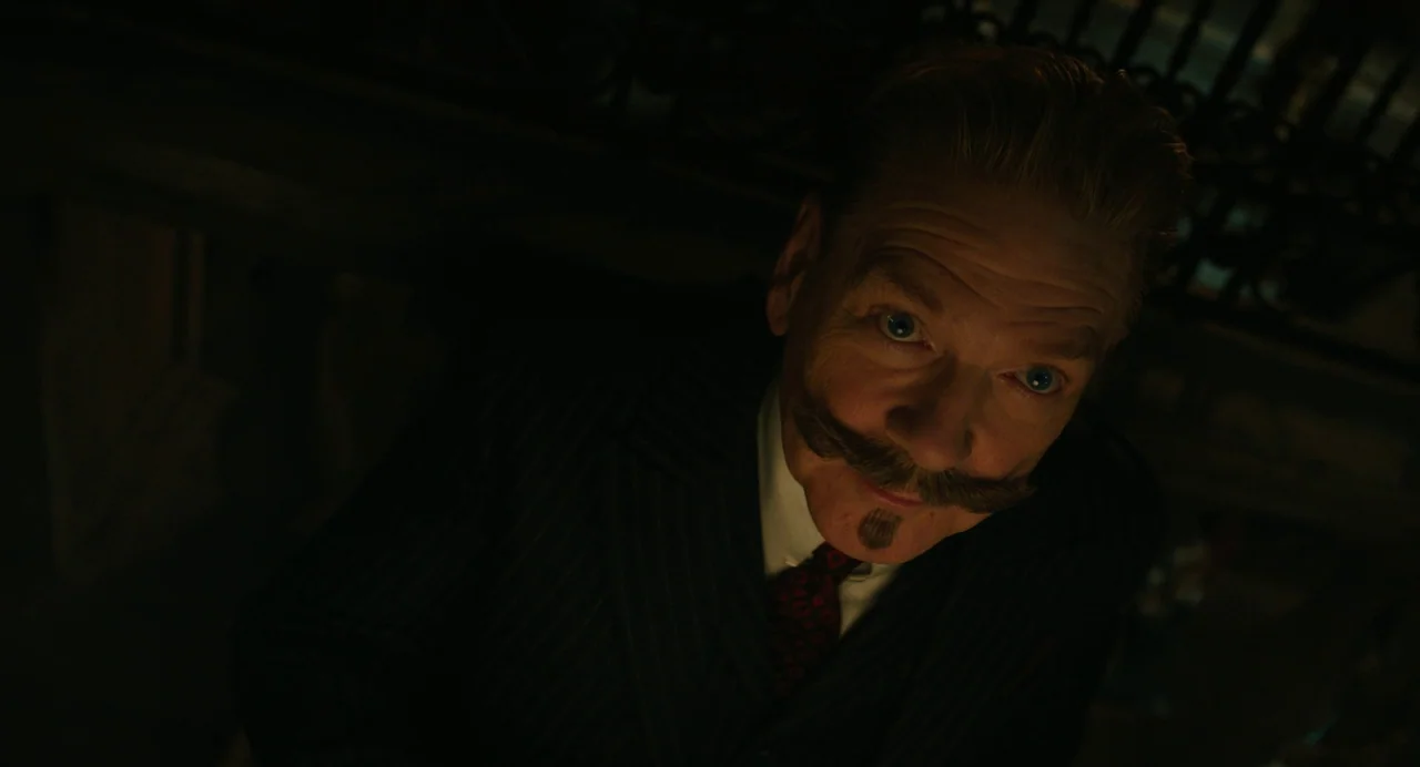 A Haunting In Venice (2023) movie still: closeup — The interior of a dimly lit, wood-paneled passenger cabin on a train. A man with a distinctive…