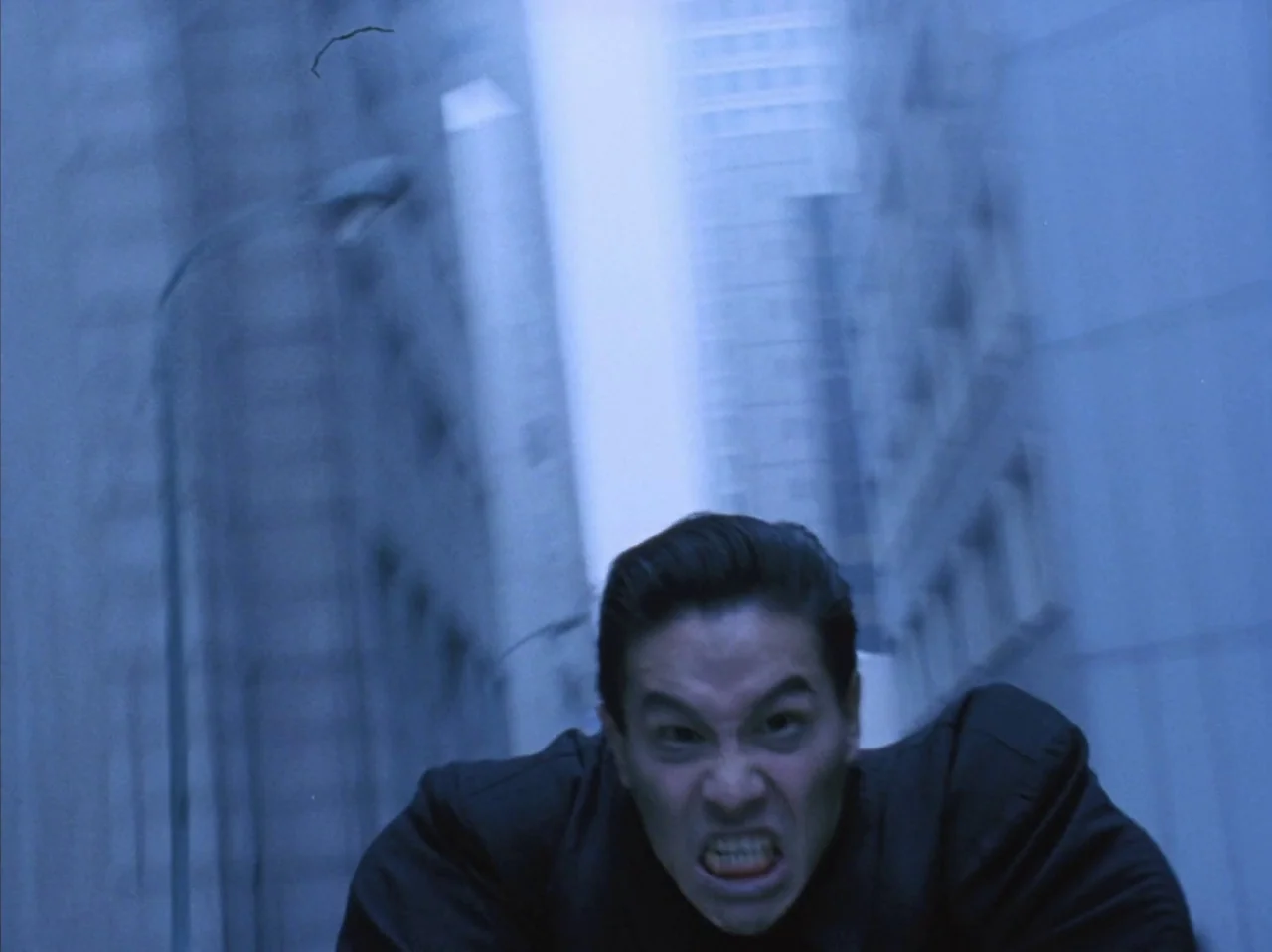 Tetsuo II: Body Hammer (1992) movie still: medium — An urban canyon at night features tall buildings flanking a narrow street space. A man in a dark…