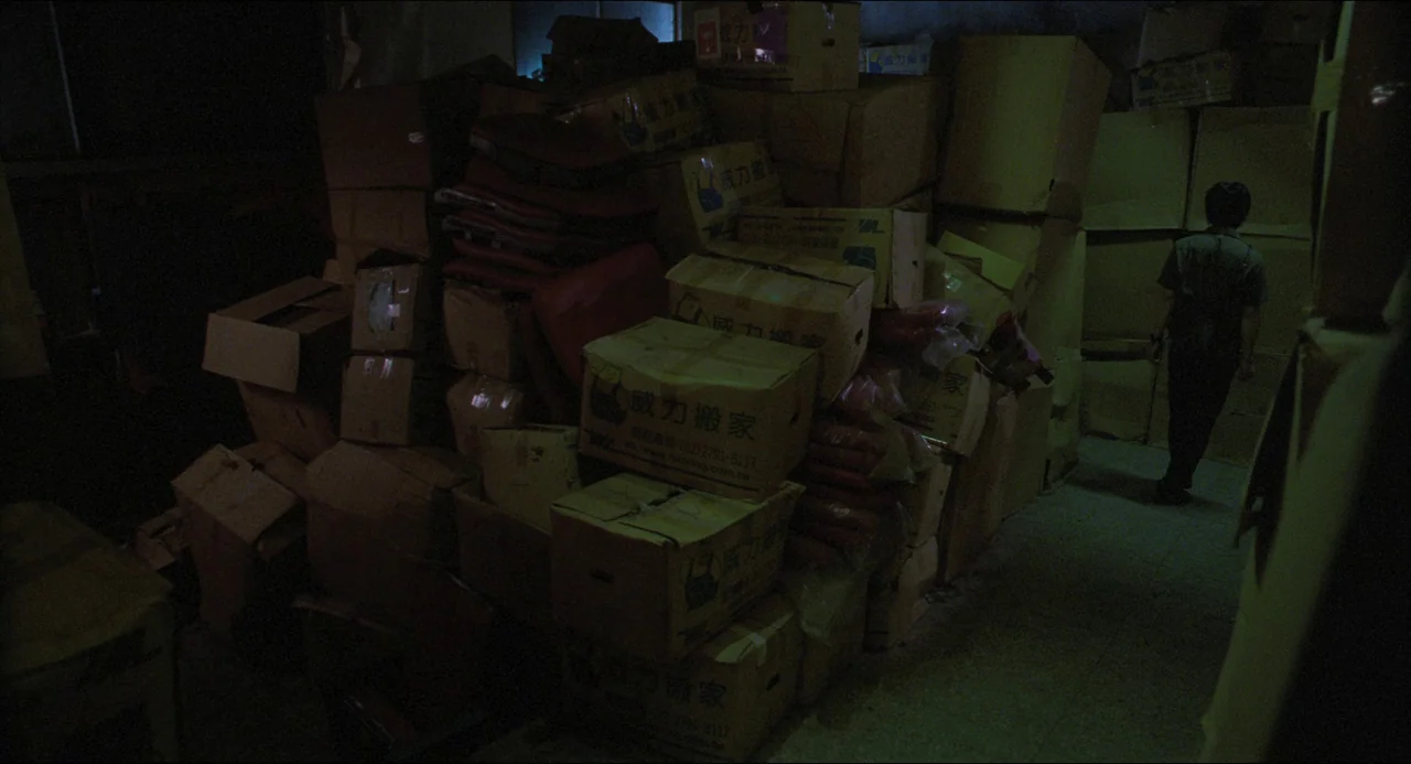 Goodbye, Dragon Inn (2003) movie still: wide — A dimly lit room is filled with uneven, high stacks of cardboard moving boxes. A lone man stands at…