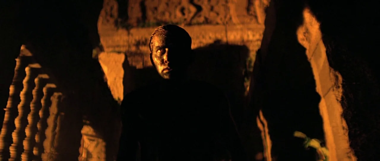 Apocalypse Now (1979) movie still: medium — An ancient stone chamber features carved reliefs visible in the background shadow. A man covered in mud…
