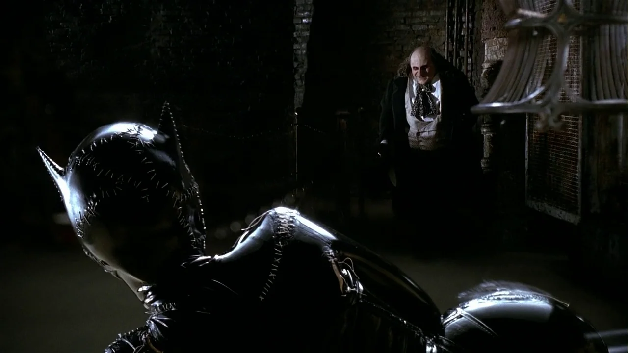 Batman Returns (1992) movie still: medium — A dark, brick-walled underground space with deep shadows obscuring the background architecture. A woman…