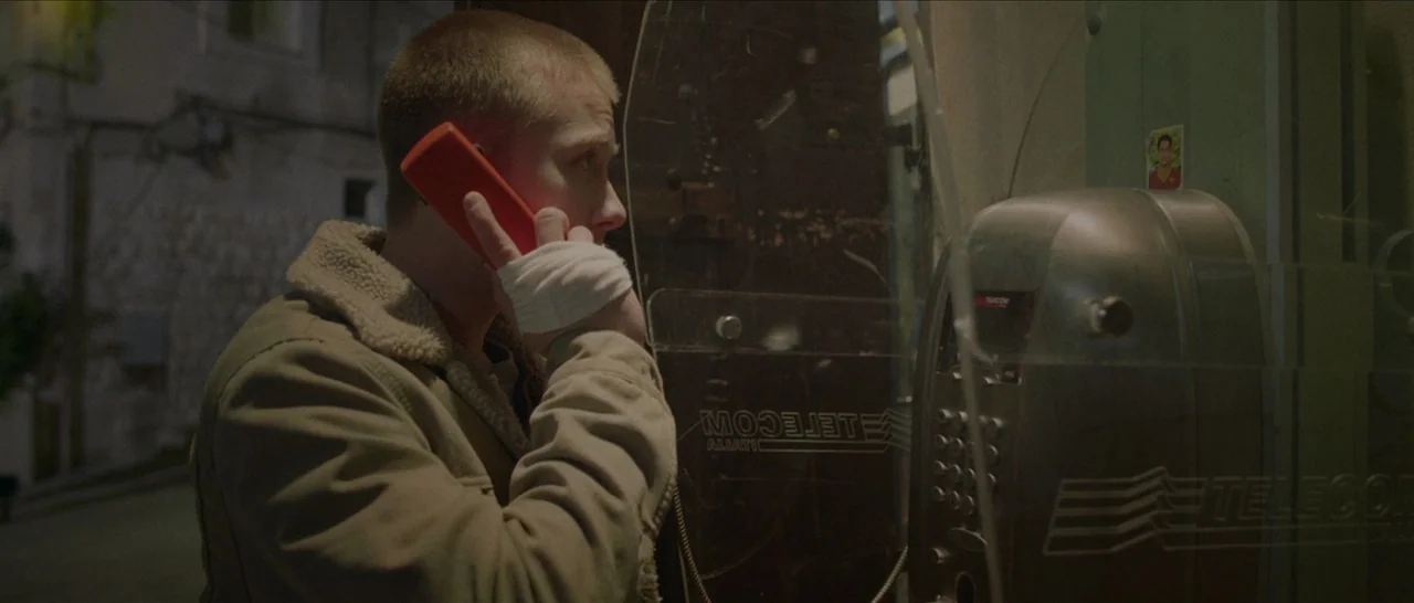 Spring (2014) movie still: medium — A man stands inside a metallic public telephone booth at night. He holds a bright orange receiver to his ear…