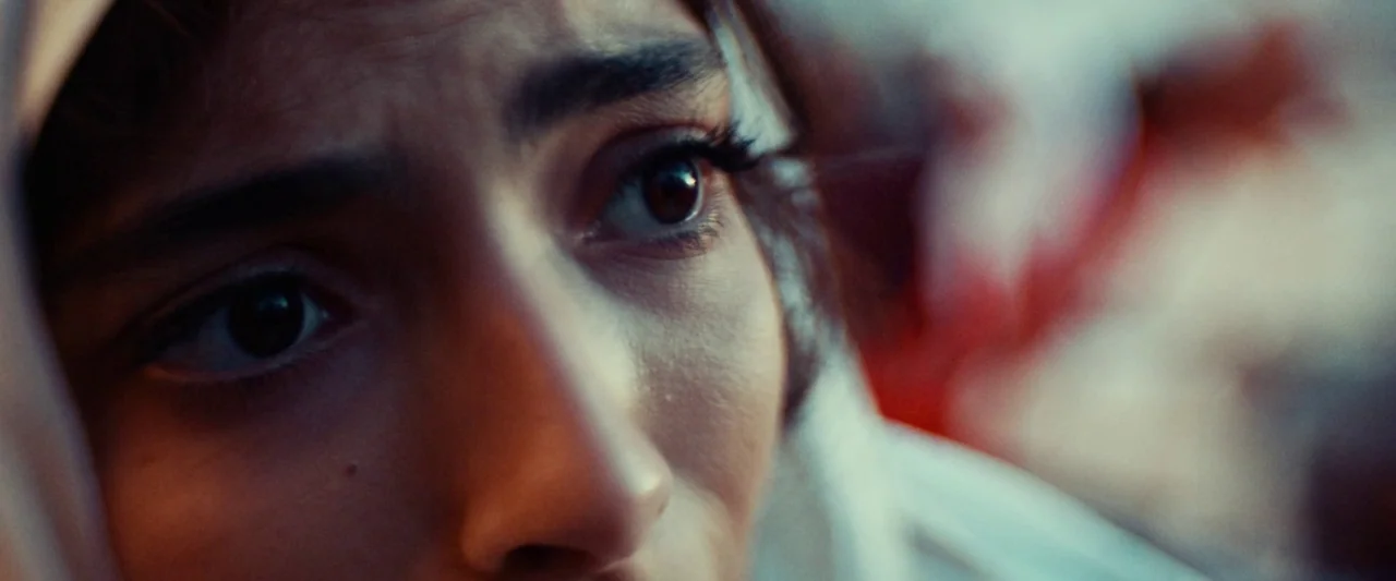 Animalia (2023) movie still: closeup — A close-up view focuses on a woman's eyes framed by a head covering. The background shows blurred red light…