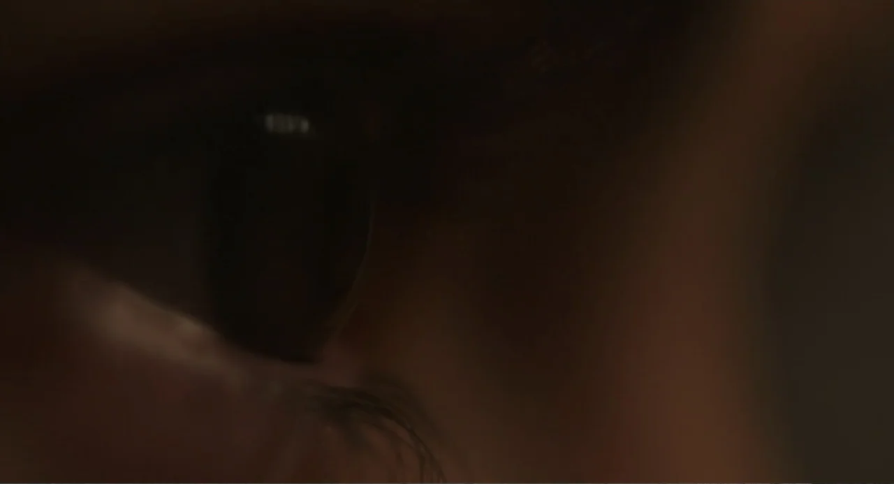Orphan: First Kill (2022) movie still: closeup — A dark interior space contains the side of a human face in shadow. A single eye is visible with…