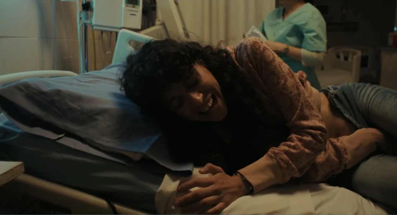 Belén (2025) movie still: medium — A hospital room contains a bed with blue linens and medical monitoring equipment. A woman lies on the bed in pain…