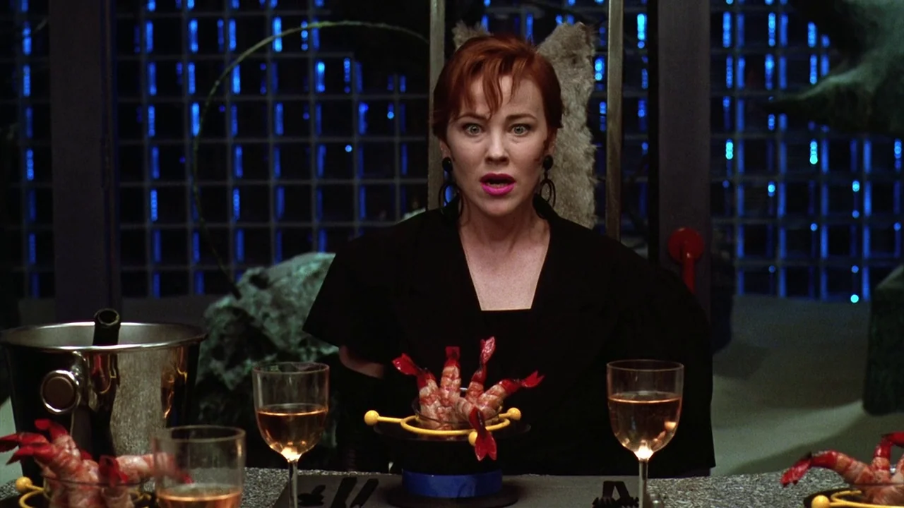 Beetlejuice (1988) movie still: medium — A dark dining room features a glass block wall illuminated by blue internal light. A woman in a black dress…