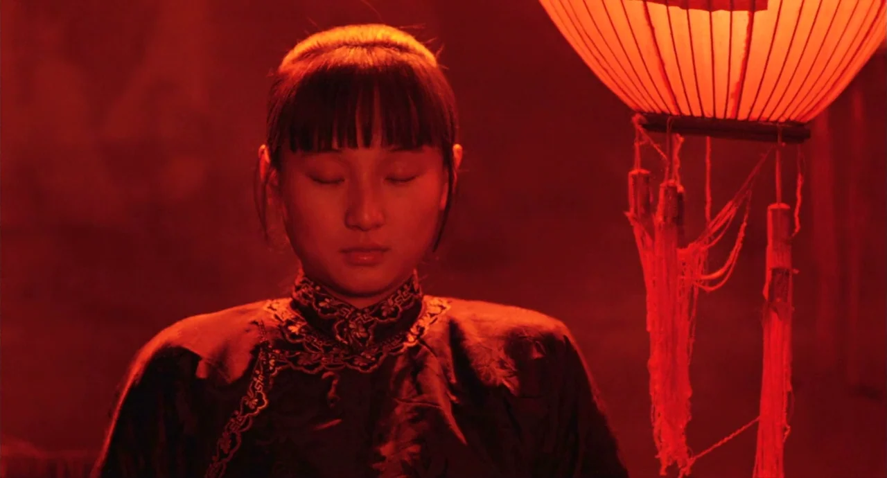 Raise The Red Lantern movie still: medium — A woman stands in a dimly lit room against a solid dark red background. She wears a black qipao with…