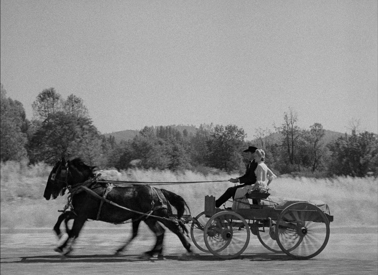 High Noon (1952) movie still: wide — A horse-drawn wagon travels across a dry field with distant hills in the background. A man and a woman sit…