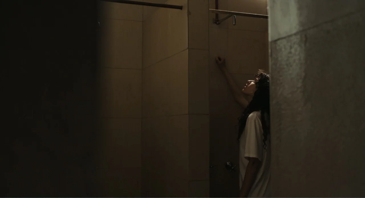 Belén (2025) movie still: medium — A tiled interior space with a metal shower head mounted on the wall. A woman stands in the stall with her hand…