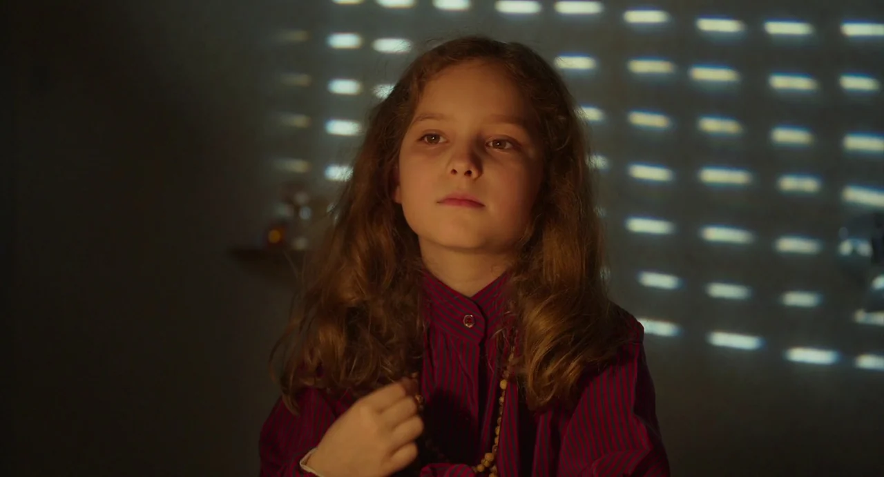 Petite Maman (2021) movie still: medium — A young girl with wavy brown hair sits in front of a wall with window blind shadows. She wears a striped…