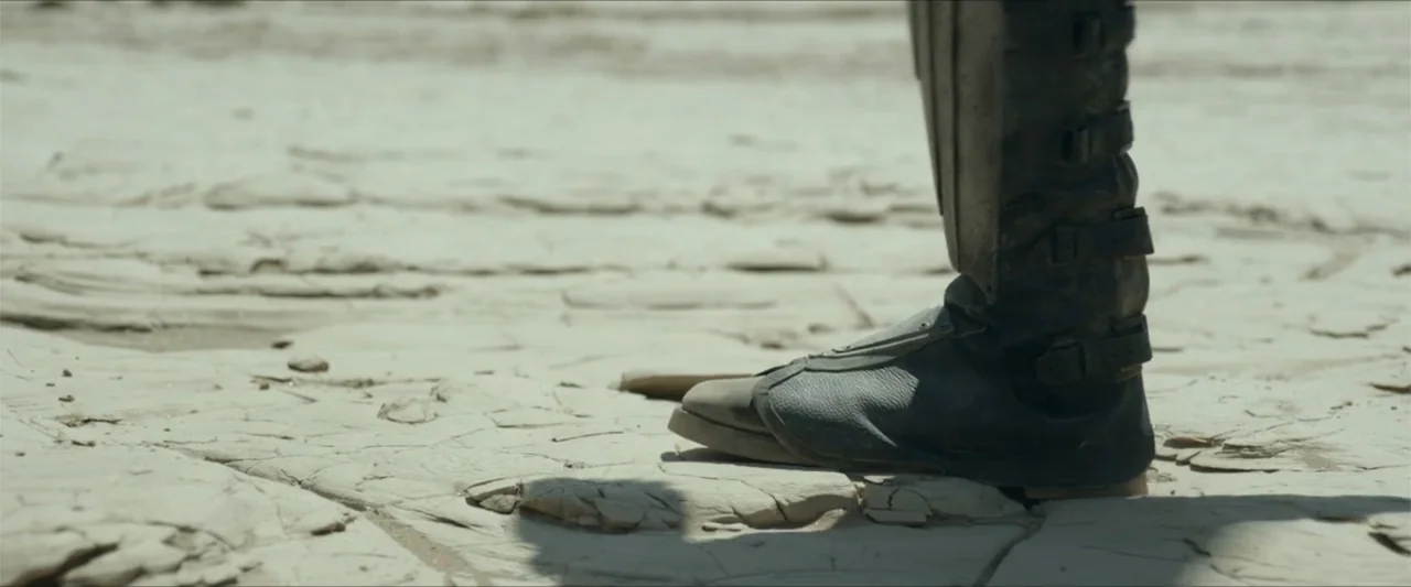 Monster Hunter (2020) movie still: closeup — A parched and cracked white earth surface stretches across the frame. A single dark armored boot stands…