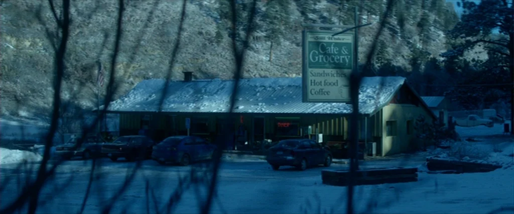 Haywire (2011) movie still: establishing — A remote roadside cafe and grocery store stands in a snowy mountain valley. Bare tree branches obscure…