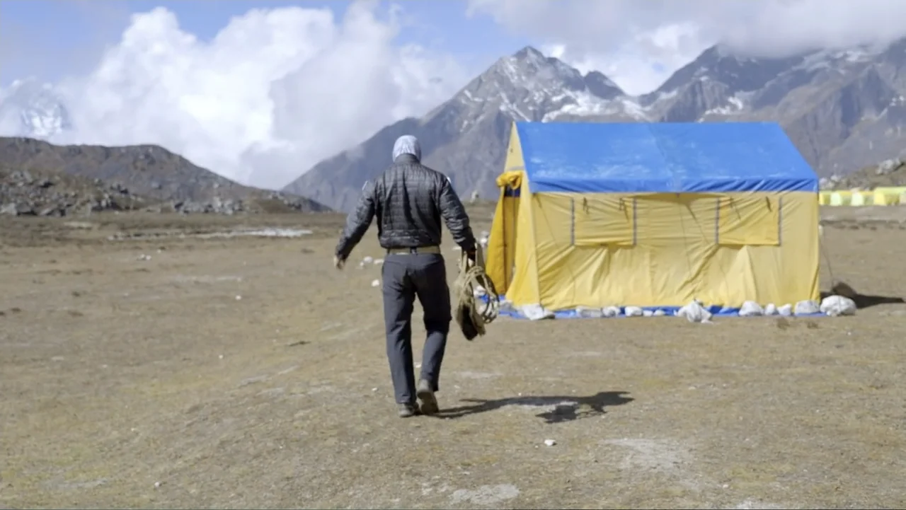 Drop Zone Everest (2023) movie still: fullbody — A high-altitude mountainous landscape features a large yellow and blue supply tent. A lone hiker…