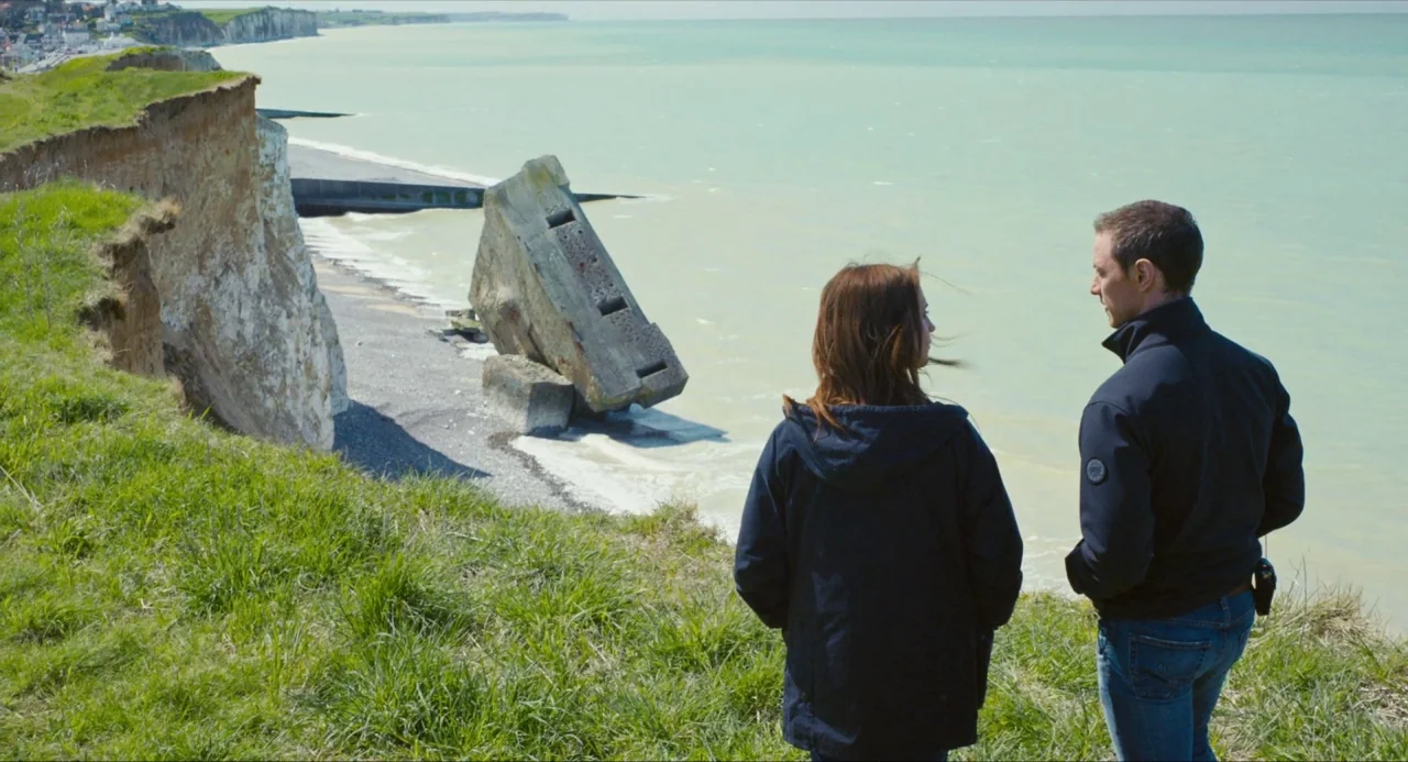 Submergence movie still: medium — A grassy cliff edge overlooks a calm bay with a concrete bunker on the beach. A man and a woman stand side by side…