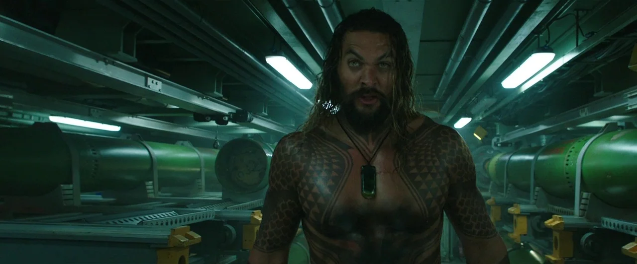 Aquaman (2018) movie still: medium — A cramped, dimly lit corridor filled with large cylindrical green weapon canisters on racks. A muscular man…