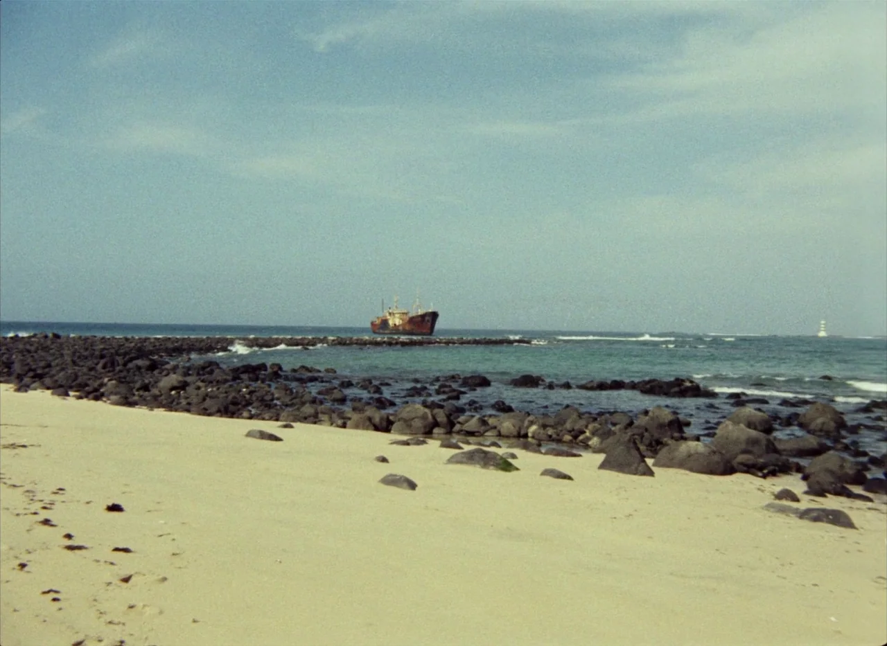 Touki Bouki (1973) movie still: wide — A sandy shoreline leads to a rocky breakwater extending into the ocean. A rusted, abandoned ship is wedged…