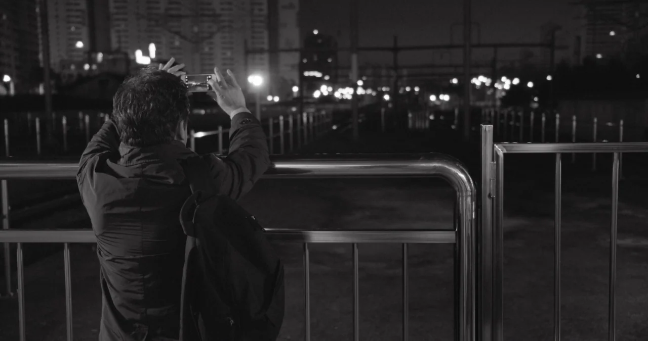 The Day After movie still: medium — A man stands behind a stainless steel railing at an urban train station at night. He holds a smartphone with…