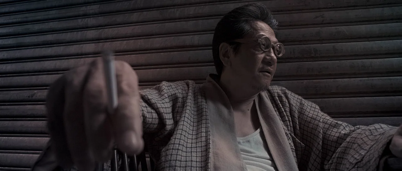 Rigor Mortis (2013) movie still: medium — An east Asian man sits before a closed metal security door at night. He holds a cigarette in his…