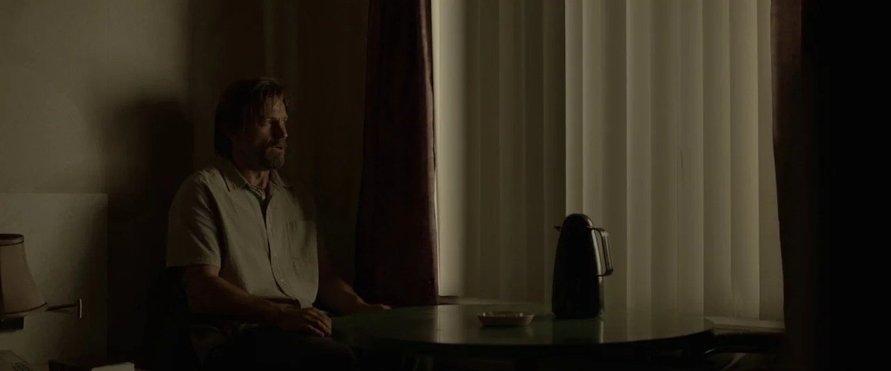 Small Crimes (2017) movie still: medium — A dimly lit room features a round table beside a window with closed blinds. A man in a light shirt sits at…