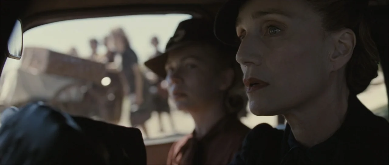 Suite Française (2015) movie still: medium — Two women wearing vintage hats sit inside the front cabin of a 1940s automobile. They look out the side…