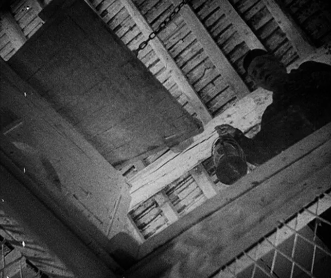 Vampyr (1932) movie still: medium — A wooden attic interior features exposed rafters and a square floor hatch. A man stands on the floor above…