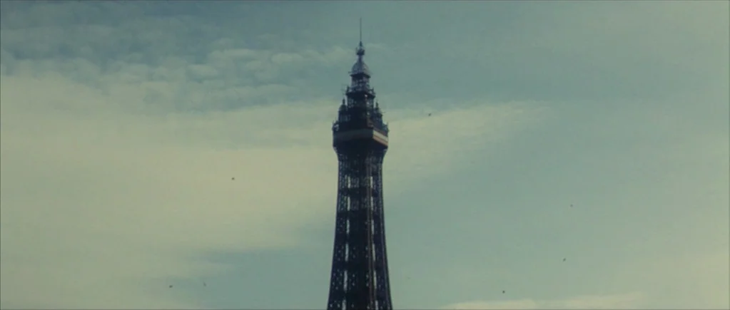 Nowhere Boy movie still: wide — A tall iron lattice tower rises vertically against a cloudy, pale blue sky. Several small birds are silhouetted in…
