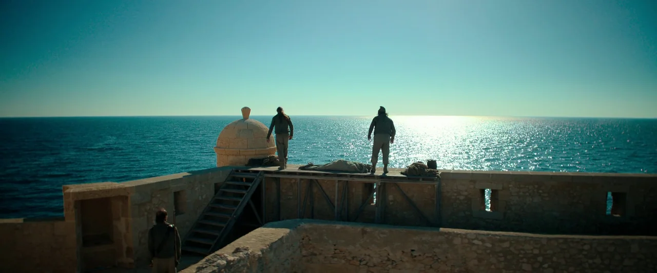The Count of Monte-Cristo (2024) movie still: wide — A stone fortification sits above a calm ocean expanse under a bright clear sky. Three men stand…