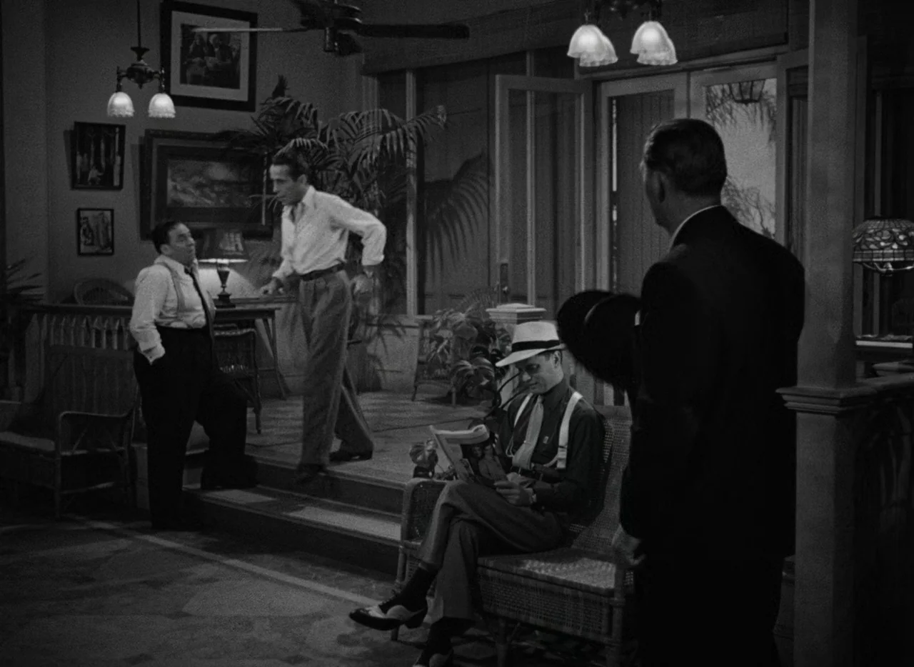 Key Largo (1948) movie still: wide — A spacious room features rattan furniture and large windows opening onto a dark exterior. Four men are present…