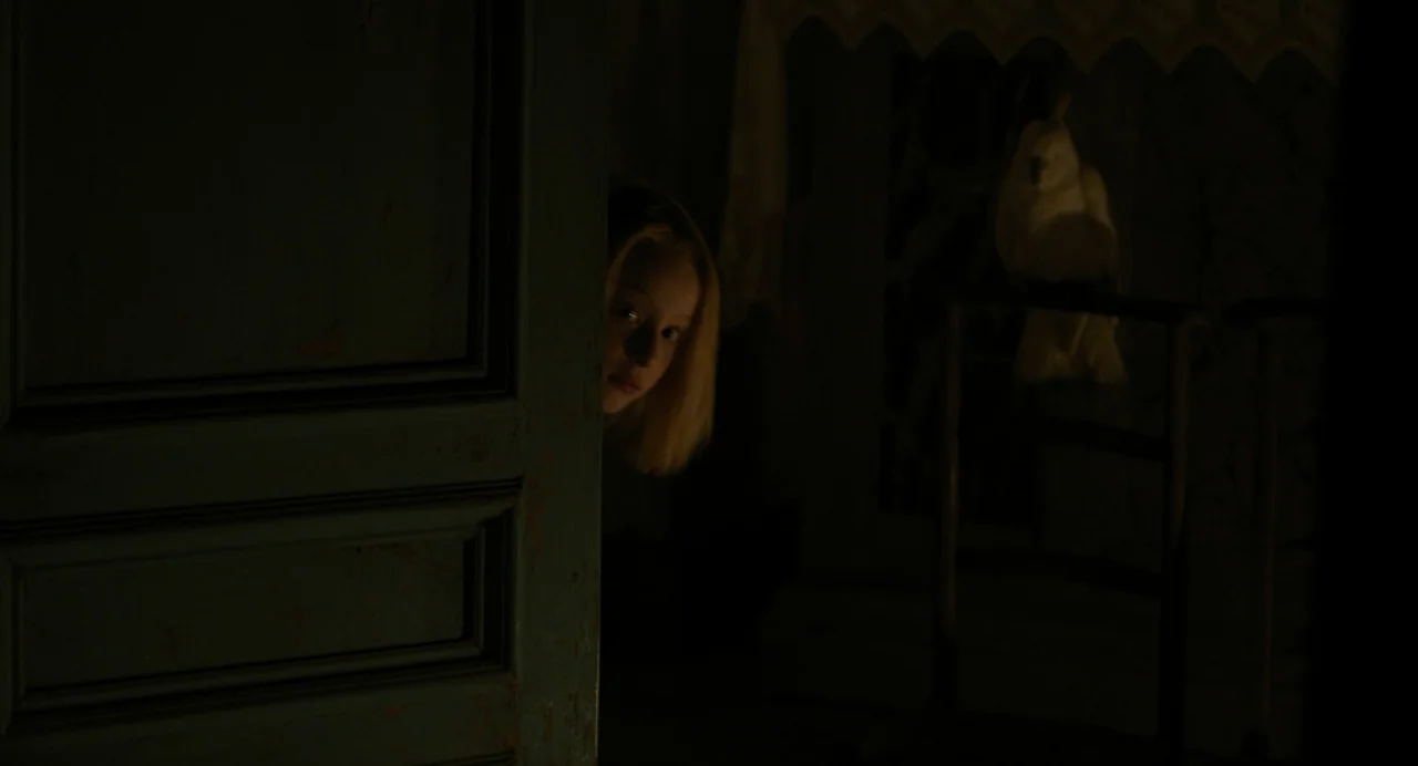 A Haunting In Venice (2023) movie still: closeup — A darkened interior scene features a doorframe partially obscuring a young girl's face. A white…