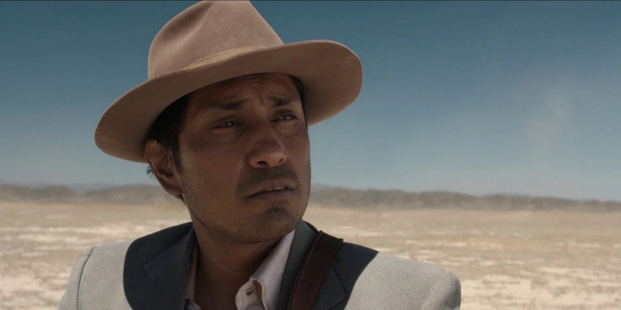 Pedro Páramo (2024) movie still: medium — An arid, sun-bleached desert landscape extends to the distant horizon under a clear sky. A man in a fedora…