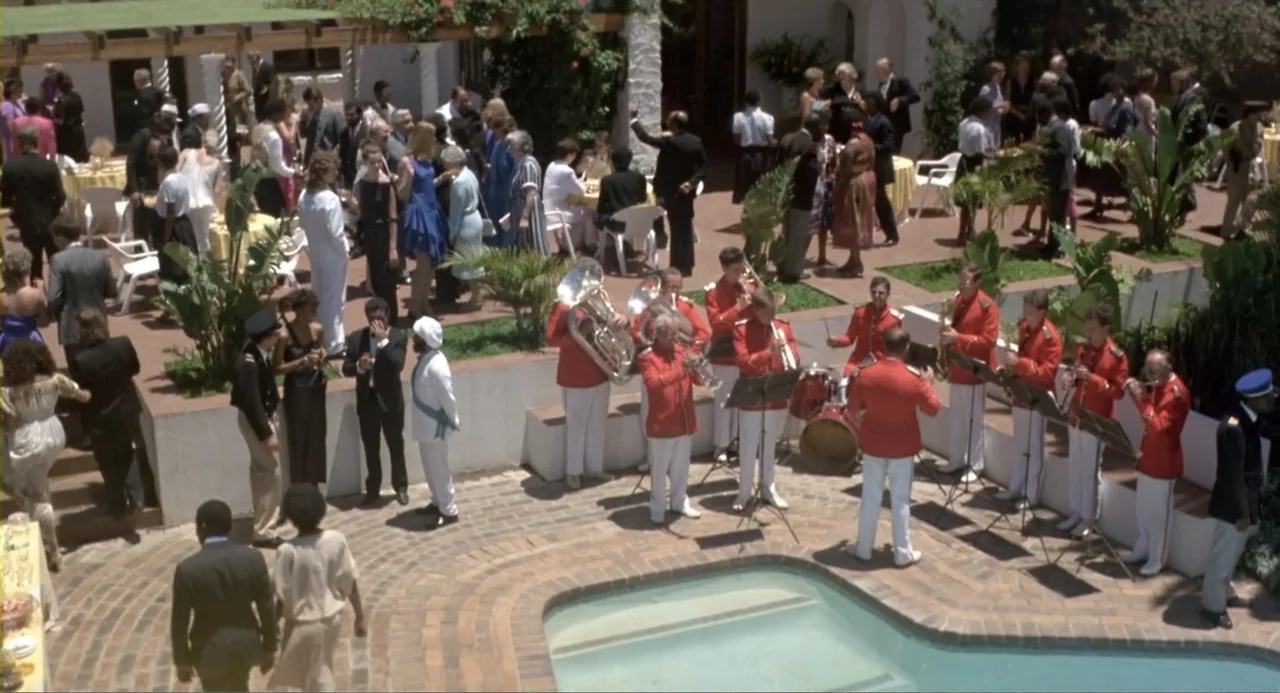American Ninja 2: The Confrontation (1987) movie still: wide — A sunlit outdoor courtyard features a paved pool area and a large social gathering. A…