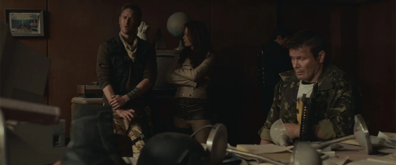 The Condemned 2 (2015) movie still: medium — A dimly lit room features wood-paneled walls and a desk covered with scattered papers. Three people in…