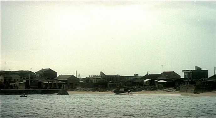 The Boys From Fengkuei (1983) movie still: establishing — A quiet coastal village sits along the shoreline under an overcast, hazy sky. Several…