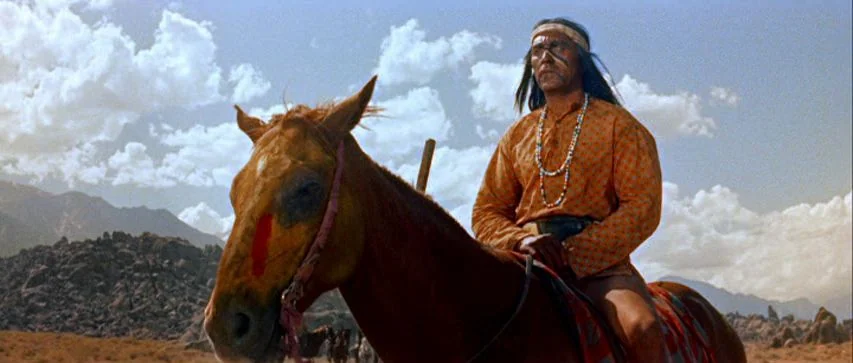 Ride Lonesome (1959) movie still: medium — A rugged desert environment with large rock formations beneath a bright, cloudy sky. A man with face…
