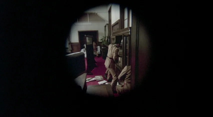 Mishima: A Life in Four Chapters (1985) movie still: wide — A dimly lit room with dark wood cabinets and a deep red carpet. A man in tan…